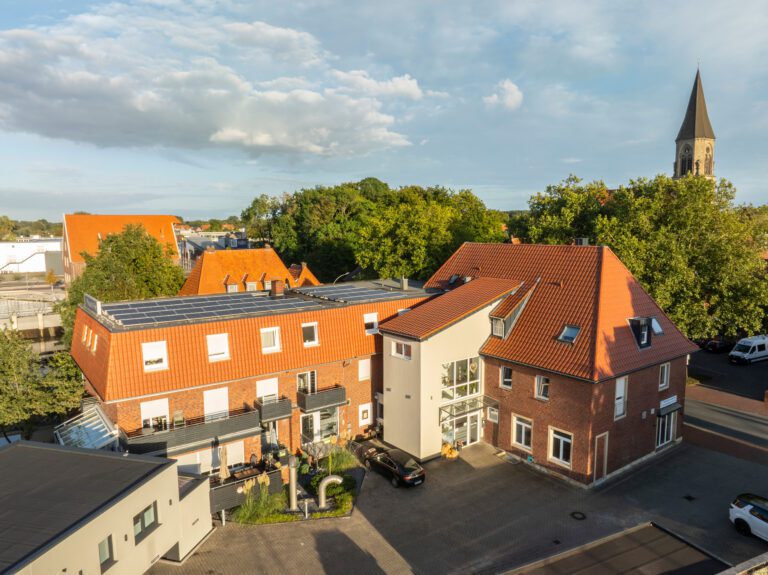 Luftaufnahme eines mehrstöckigen Backsteingebäudes mit roten Dächern und Sonnenkollektoren, umgeben von Bäumen und anderen Gebäuden. Im Hintergrund steht eine Kirche mit einem hohen Kirchturm unter einem teilweise bewölkten Himmel.
