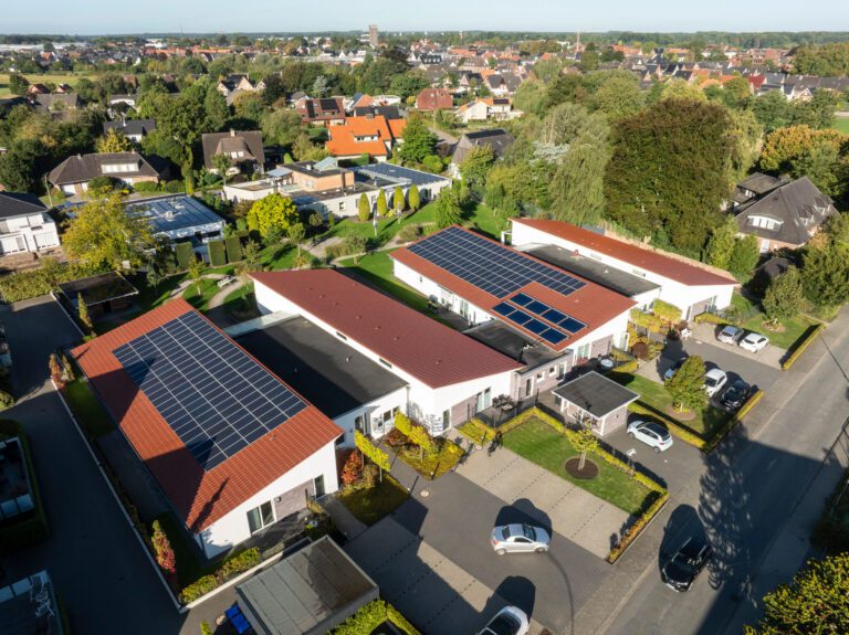 Luftaufnahme eines Wohnviertels mit modernen, einstöckigen Häusern mit roten Dächern und Sonnenkollektoren. Das Gebiet ist von Bäumen, Grünflächen und geparkten Autos umgeben, mit weiteren Häusern und Gebäuden im Hintergrund.