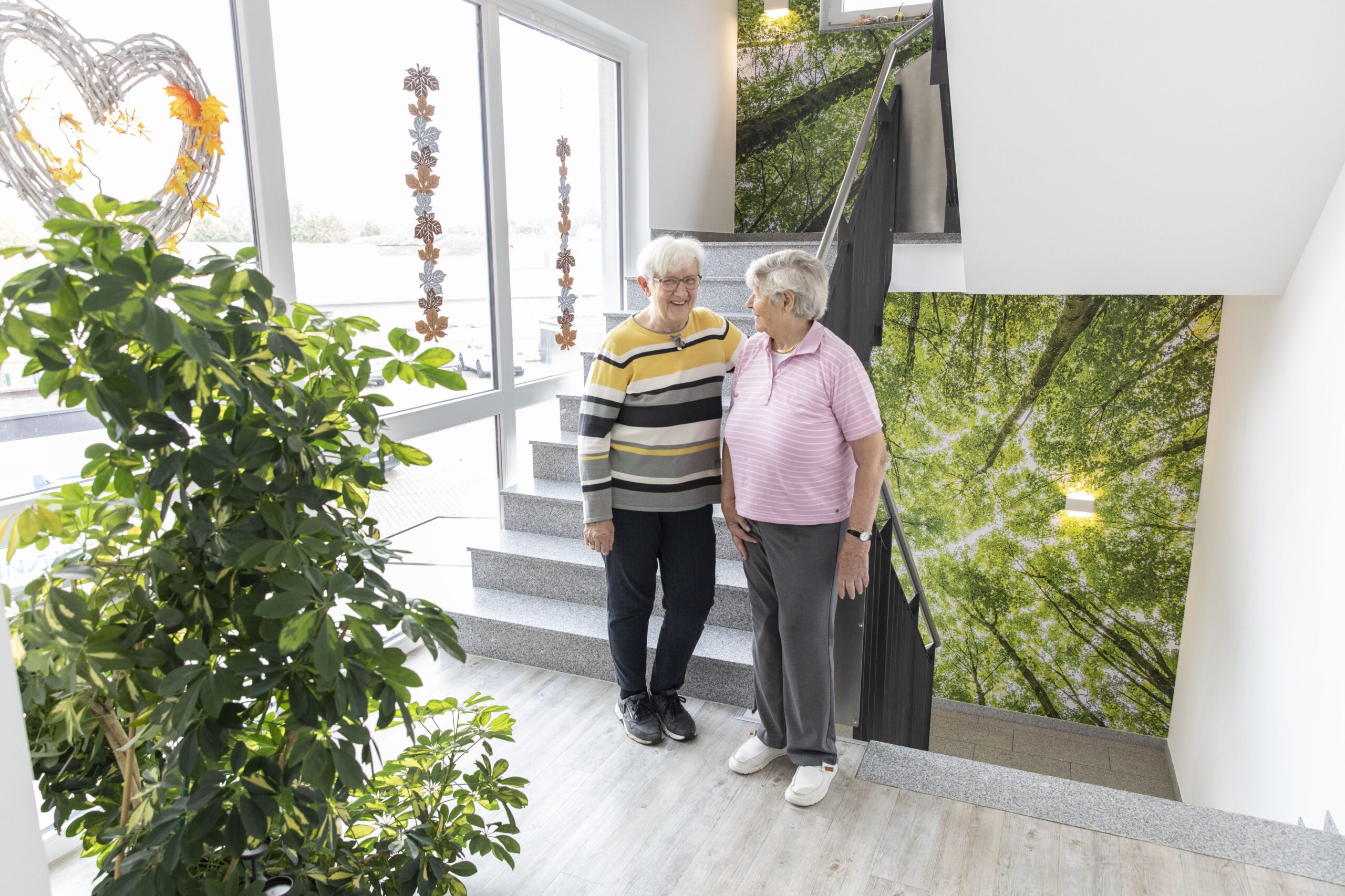 Zwei ältere Menschen stehen lächelnd auf einer hell erleuchteten Treppe mit großen Fenstern und grüner Wandmalerei; eine Topfpflanze steht in der Nähe und schafft eine fröhliche und einladende Atmosphäre.