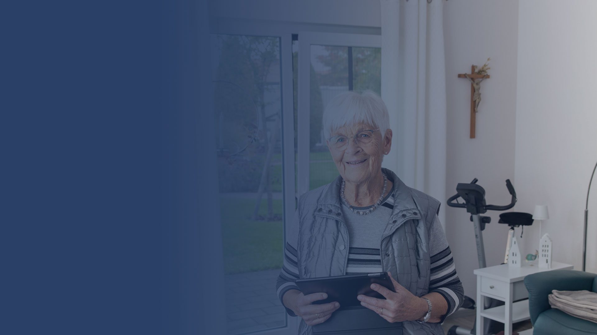 Eine ältere Frau mit kurzen weißen Haaren und Brille steht in einem hellen Raum, lächelt und hält ein Tablet in der Hand. Hinter ihr befinden sich ein Fenster, ein Fitnessgerät, ein Nachttisch und ein Kreuz an der Wand.