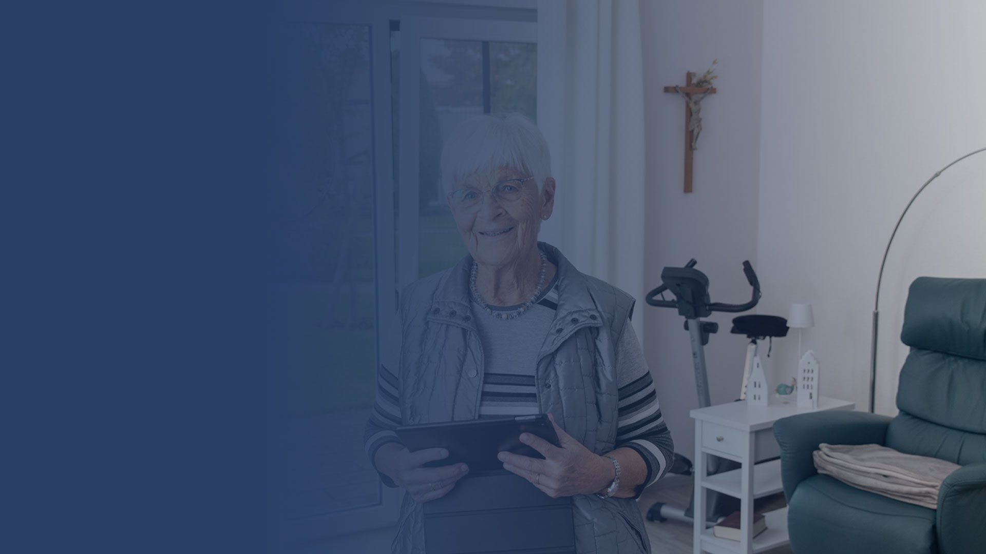 Eine ältere Frau mit kurzen weißen Haaren steht in einem Wohnzimmer, lächelt und hält ein Tablet in der Hand. Hinter ihr stehen ein stationäres Fahrrad, ein grüner Stuhl und ein an der Wand befestigtes Kruzifix. Die linke Seite des Bildes ist stark blau schattiert.