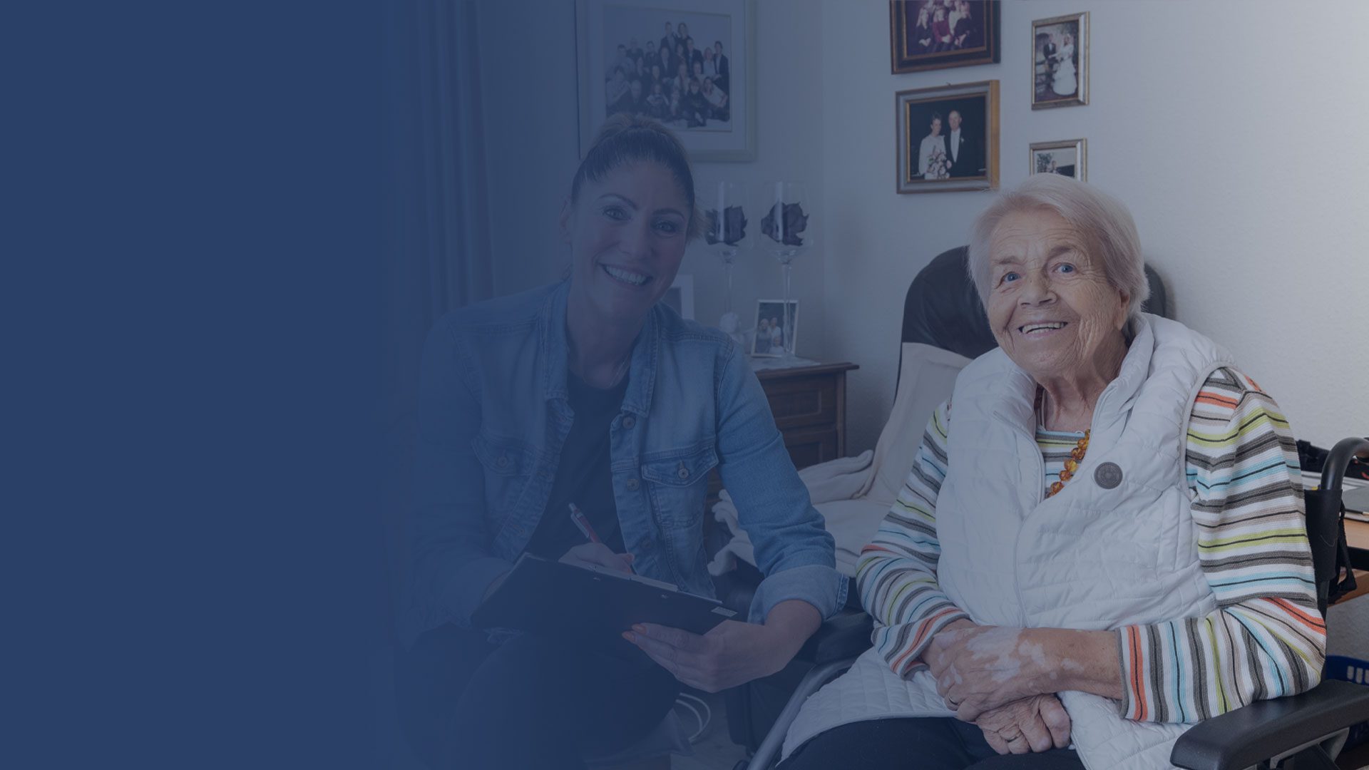 Eine lächelnde ältere Frau im Rollstuhl sitzt neben einer jüngeren Frau, die ein Klemmbrett hält. Sie befinden sich in einem gemütlichen Raum mit Familienfotos an der Wand. Die linke Seite des Bildes hat eine blaue Überlagerung.