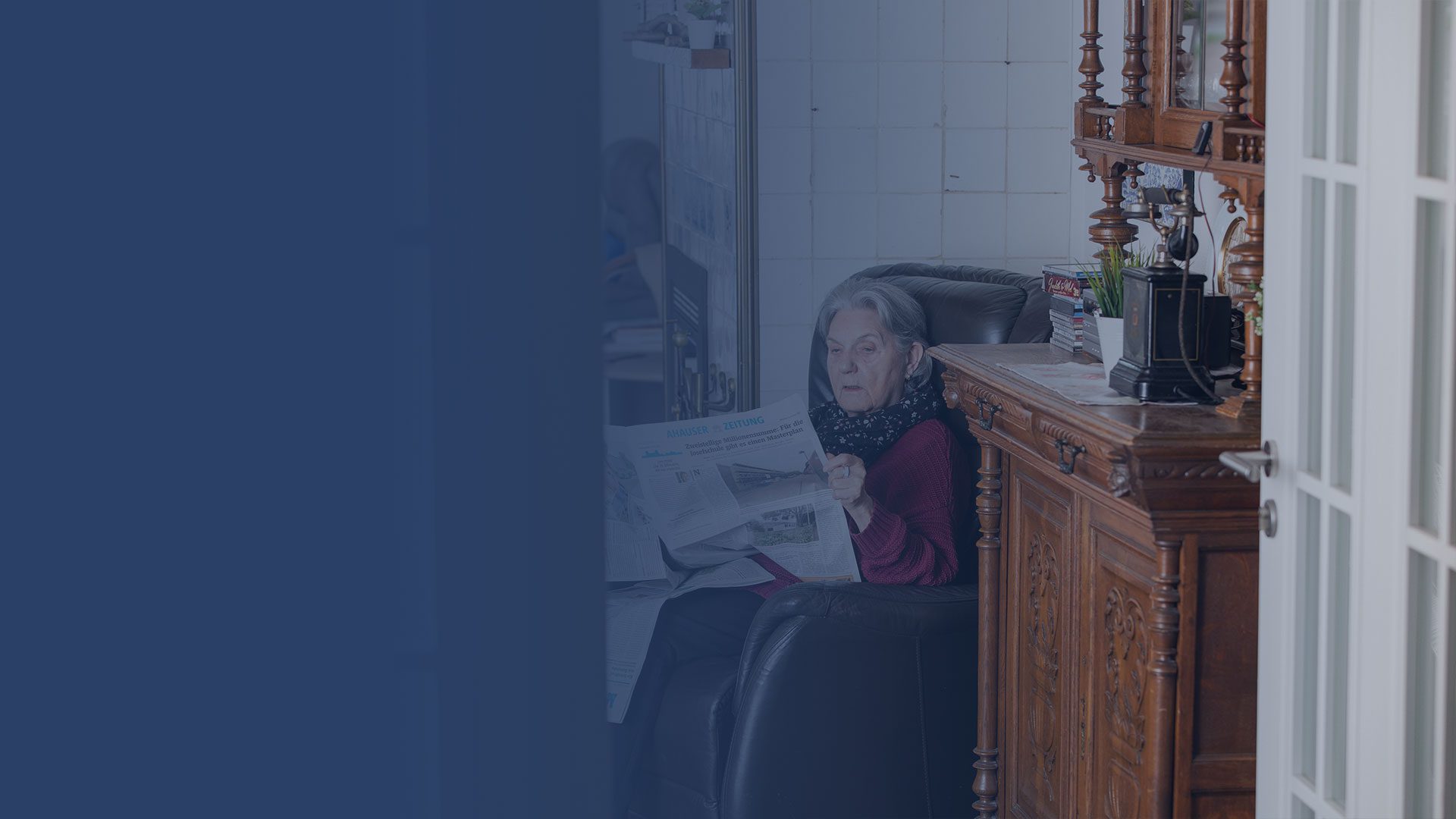 Eine ältere Frau mit grauem Haar sitzt in einem Sessel, hält eine Zeitung in der Hand und gestikuliert mit der Hand in einem Raum mit weiß gefliesten Wänden und einem Holzschrank. Die linke Seite des Bildes ist von einem blauen Overlay überlagert.