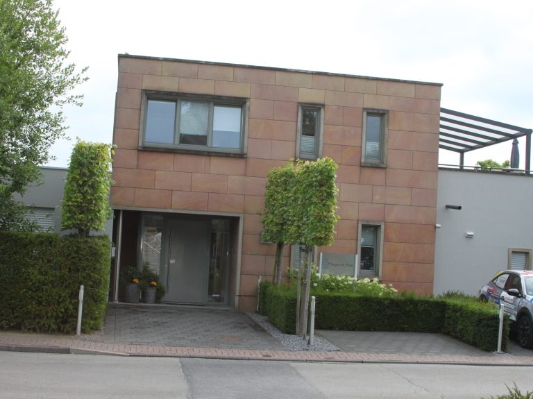 Ein modernes zweistöckiges Haus mit rechteckigem Grundriss und hellbrauner Steinfassade, großen Fenstern, beschnittenen Büschen und einem geparkten Auto, das teilweise auf der rechten Seite der Einfahrt zu sehen ist.