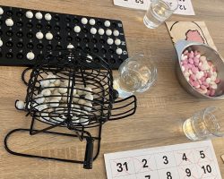 Bingo-Nachmittag in der Tagespflege mit Plan: Bingo-Trommel und Zahlenbrett auf einem Holztisch, daneben ausgefüllte Bingokarten, Wassergläser und eine Schale mit Süßigkeiten.