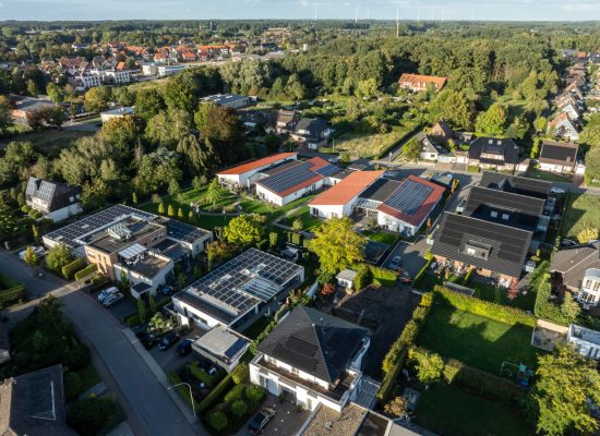 Luftaufnahme eines Vorstadtviertels mit Häusern, auf deren Dächern Sonnenkollektoren angebracht sind, umgeben von Bäumen und Grünflächen, mit einer Stadt und einem Wald im Hintergrund unter blauem Himmel.