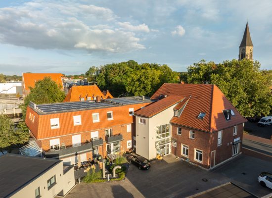 Luftaufnahme eines mehrstöckigen Backsteingebäudes mit roten Dächern und Sonnenkollektoren, umgeben von Bäumen und anderen Gebäuden. Im Hintergrund steht eine Kirche mit einem hohen Kirchturm unter einem teilweise bewölkten Himmel.