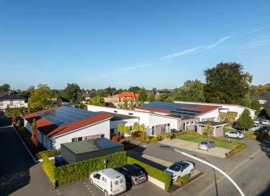 Luftaufnahme von modernen Häusern mit Sonnenkollektoren auf den Dächern, umgeben von Grünflächen und parkenden Autos an einem sonnigen Tag.
