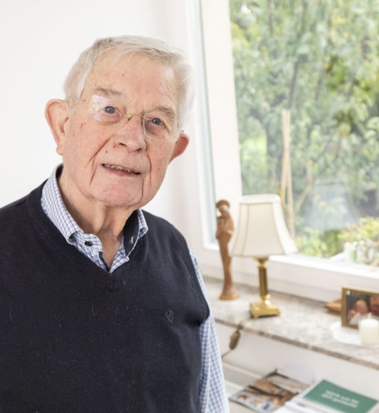 Ein älterer Mann mit weißem Haar und Brille, der ein kariertes Hemd und eine dunkle Pulloverweste trägt, steht in einem Innenraum an einem Fenster, durch das Sonnenlicht fällt; auf der Fensterbank hinter ihm sind Fotos und Dekoration zu sehen.