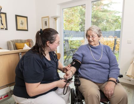 Eine Pflegekraft prüft den Blutdruck einer älteren Frau im Rollstuhl in einem gemütlichen, gut beleuchteten Raum mit einem großen Fenster, aus dem man die Herbstbäume sehen kann.