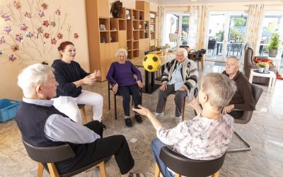 Sechs ältere Menschen und eine jüngere Frau sitzen in einem hellen Raum im Kreis, lächeln und werfen sich gegenseitig einen gelb-schwarzen Ball zu, was an eine Gruppenaktivität oder Übung in einem Pflegeheim oder Gemeindezentrum denken lässt.