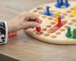 Eine ältere Hand bewegt eine rote Spielfigur auf einem hölzernen Brettspiel mit bunten Figuren, das auf einem Holztisch gespielt wird.