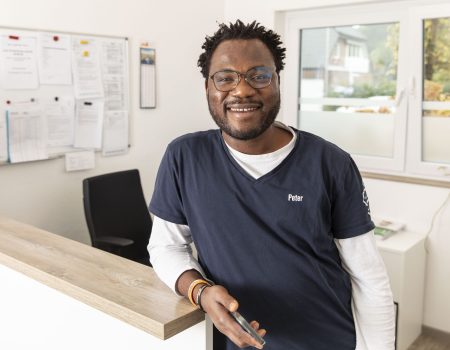 Ein lächelnder Mann mit Brille und marineblauem Kittel namens Peter steht an der Rezeption in einem hellen Büro und hält ein Smartphone in der Hand. Im Hintergrund sind ein schwarzes Brett und ein Fenster mit Blick nach draußen zu sehen.