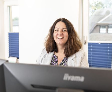 Eine Frau mit schulterlangem braunem Haar, die einen hellgrauen Blazer und ein geblümtes Oberteil trägt, lächelt, während sie an einem Schreibtisch in einem Büro mit großen Fenstern und blauen Jalousien sitzt.