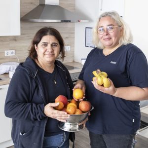 Zwei Frauen stehen in einer modernen Küche; die eine hält eine Schale mit Äpfeln, die andere ein Bündel Bananen. Beide lächeln und tragen dunkelblaue Hemden.