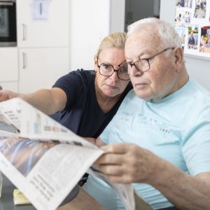 Eine Frau sitzt neben einem älteren Mann im Rollstuhl, beide schauen in eine Zeitung. Die Frau zeigt auf etwas in der Zeitung und hilft dem Mann beim Lesen. Sie befinden sich in einer hellen Küche mit Fotos an der Wand hinter ihnen.