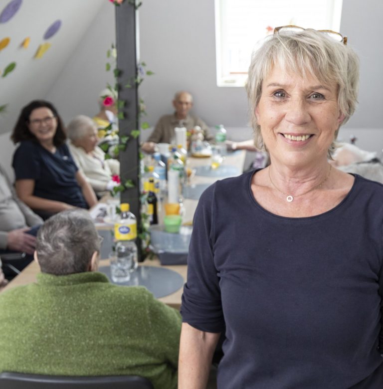 Eine lächelnde Frau steht im Vordergrund eines hellen Raums, während im Hintergrund mehrere ältere Erwachsene an einem Tisch sitzen und sich bei einem Getränk unterhalten.