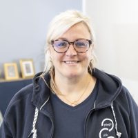 Eine lächelnde Frau mit blondem Haar und Brille trägt eine dunkle Jacke und steht in einem Raum. Hinter ihr stehen eine Couch mit Bilderrahmen und ein an der Wand montierter Fernseher.