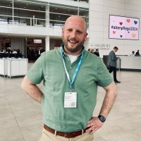 Ein lächelnder Mann in einem grünen Polohemd mit einem Schlüsselband steht in einem modernen Konferenzzentrum. Hinter ihm stehen Menschen, Theken und ein Schild mit der Aufschrift #altenpflege2024 und bunten Herzen.